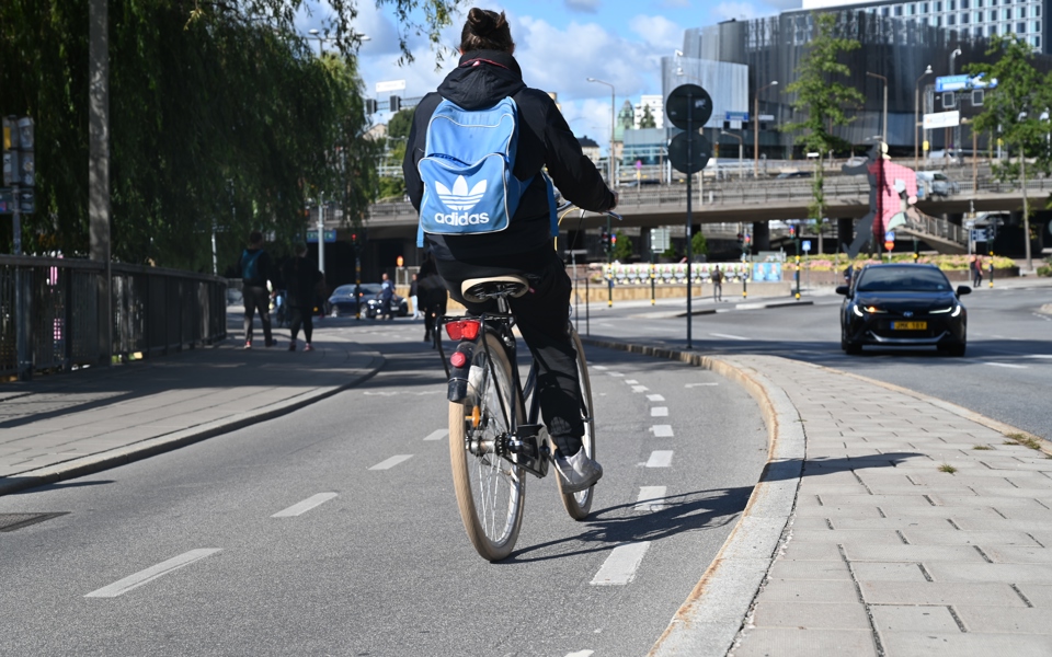 Cyklande person med ryggsäck sedd bakifrån på en cykelbana. I bakgrunden syns en bil, byggnader och människor i rörelse, fotografi.
